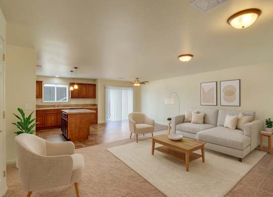 A bright open-concept living room here at Phoenix Ridge featuring a neutral sofa with accent pillows, a wooden coffee table, cozy upholstered chairs, and simple wall art, flowing into a kitchen with warm wood cabinetry, an island, tiled flooring, and sliding glass doors that bring in natural light.