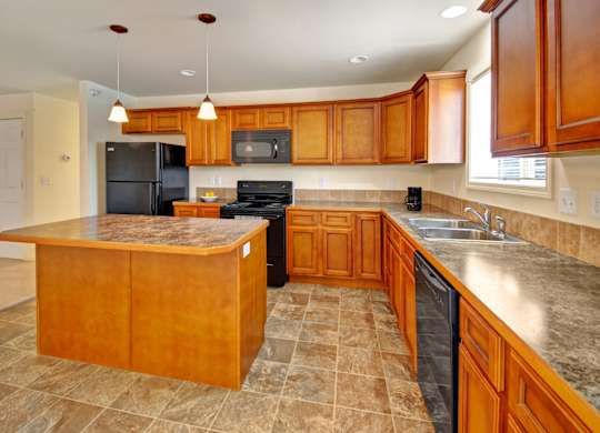 A warm modern kitchen here at Phoenix Ridge featuring rich wood cabinetry, a spacious center island with a stone-look countertop, black appliances, pendant lighting, and durable tile flooring, offering an open layout with ample counter space and natural light from a nearby window.
