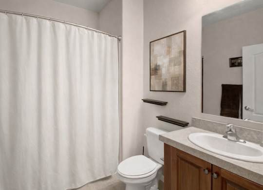 A clean bathroom here at Prairie Vista featuring a single-sink vanity with a light countertop, wood cabinetry, a large wall mirror, a toilet, floating wall shelves, neutral wall art, and a shower-tub combo with a white curtain and light tile flooring.