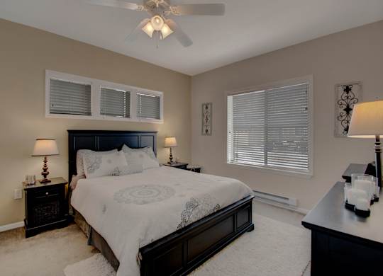 A cozy bedroom here at Prairie Vista featuring a dark wood bed with light patterned bedding, matching nightstands with table lamps, a ceiling fan with light, soft carpet flooring, neutral walls, decorative wall accents, a dresser, and windows with white blinds allowing natural light into the space.
