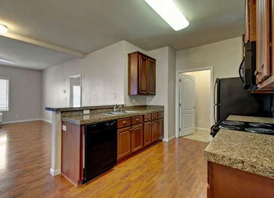 A bright open kitchen here at Prairie Vista featuring warm wood cabinetry, granite-style countertops, a double sink with breakfast bar seating, black appliances including a dishwasher and range, wood-look flooring, overhead lighting, and an open view into the living area with large windows bringing in natural light.