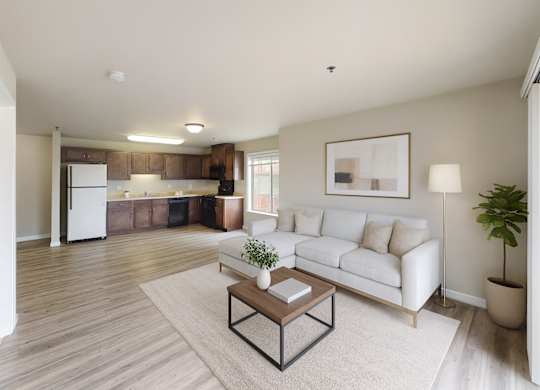 A bright, modern living room here at Renaissance Heights featuring light wood-style flooring, a neutral sectional sofa with plush pillows, a simple coffee table on a soft area rug, abstract wall art, a floor lamp, indoor greenery, and an open kitchen with dark cabinets and white appliances in the background.