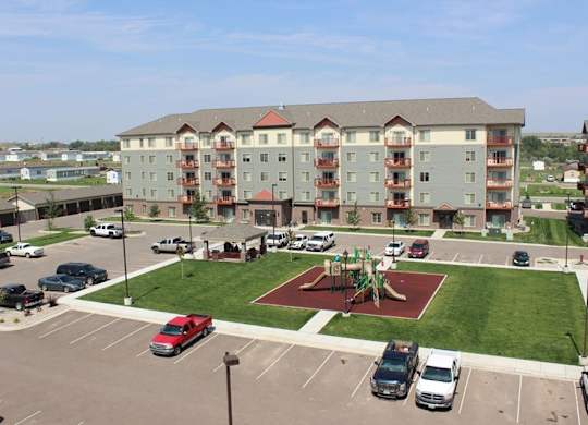 A bright aerial exterior view here at Renaissance Heights showcasing a multi-story apartment community with neutral siding, red-accented balconies, landscaped green lawns, a central playground area, a covered pavilion, and surrounding parking spaces under a clear blue sky.