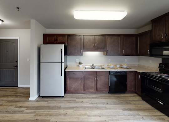 A spacious, modern kitchen here at Renaissance Heights featuring warm wood-style flooring, dark wood cabinetry, light countertops, a stainless steel refrigerator, black appliances, a double sink, and ample counter space with soft overhead lighting that creates a clean and functional cooking area.