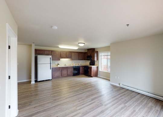 A spacious open-concept living room here at Renaissance Heights featuring wide plank wood-style flooring, neutral painted walls, and a bright layout that flows into a modern kitchen with dark wood cabinetry, light countertops, full-size appliances, and a large window that brings in natural light.