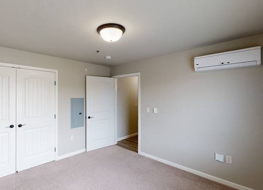 A bright and comfortable bedroom here at Renaissance Heights featuring soft neutral walls, plush carpet flooring, double white closet doors, a ceiling-mounted light fixture, and a wall-mounted mini-split unit, with an open doorway leading toward the living area for a clean and functional layout.