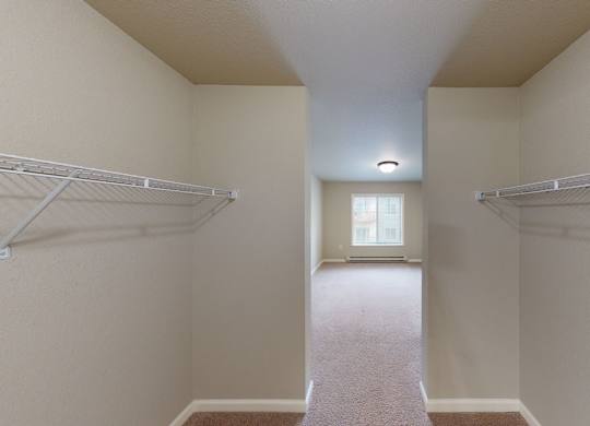 A spacious walk-in closet here at Renaissance Heights featuring dual white wire shelving on both sides, neutral walls, plush carpet flooring, and a hallway-style layout that opens toward a bright bedroom with a window and ceiling light.
