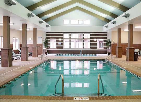 A bright indoor swimming pool here at Renaissance Heights featuring clear turquoise water, stainless steel handrails, a vaulted ceiling with exposed beams, large windows letting in natural light, tiled pool decking, and surrounding seating areas that create a relaxing, resort-style atmosphere.