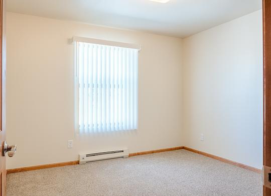 A bright, empty bedroom here at Rockridge Apartments with beige carpet, soft neutral walls, and a large window with vertical blinds that bring in natural light. Wood trim lines the baseboards, and the open layout offers flexible space for bedroom furniture while maintaining a clean, simple, and inviting feel.