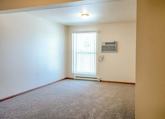 A spacious, empty living area here at Rockridge Apartments with beige carpet, neutral walls, and wood-trim baseboards. A large window with vertical blinds allows natural light to brighten the room, while a wall-mounted air conditioning unit and simple ceiling light provide added comfort in this open, flexible space.