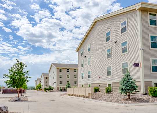 A well-maintained apartment community exterior here at Rockridge Apartments featuring beige multi-story buildings with white trim and multiple windows. A paved drive runs through the property, lined with mailboxes, landscaped rock beds, and small trees, creating an open, orderly setting under a bright, partly cloudy sky.