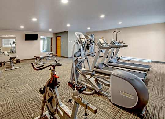 A well-equipped fitness center here at Vue 28 Apartments featuring cardio machines, weight equipment, and exercise benches arranged across a spacious, open floor plan. The room includes mirrored walls, wall-mounted televisions, patterned flooring, and bright overhead lighting, creating a comfortable and functional space for daily workouts.