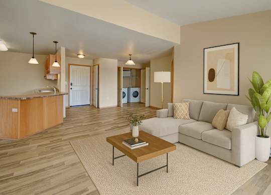 A bright, open living room here at Williston Garden Apartments with wood-style flooring, soft neutral walls, and an inviting layout that flows into the kitchen area. A light-colored sectional sofa sits on an area rug with a wooden coffee table, complemented by pendant lighting, simple wall art, and an in-home laundry area visible in the background.