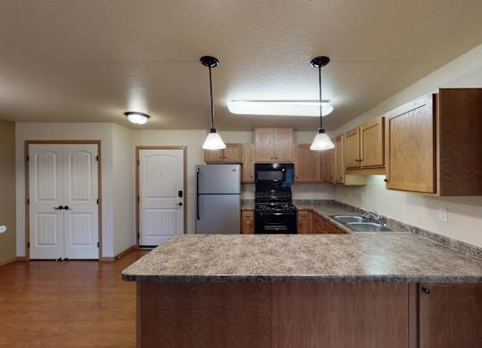 A spacious kitchen here at Williston Garden Apartments with warm wood cabinets, wood-style flooring, and expansive countertops that include a large island for added prep and seating space. The kitchen features a refrigerator, stove, microwave, and double sink, with pendant lighting and an open layout that connects easily to the surrounding living area.