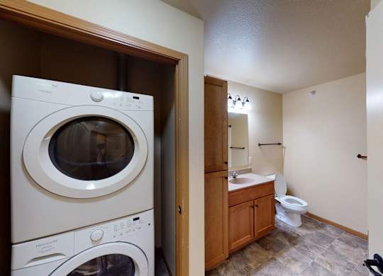 A convenient in-home laundry and bathroom area here at Williston Garden Apartments featuring a stacked washer and dryer tucked into a closet beside a bathroom vanity. The bathroom includes a wooden cabinet with sink, a white toilet, tiled flooring, and overhead lighting, offering a functional layout with everyday essentials close at hand.