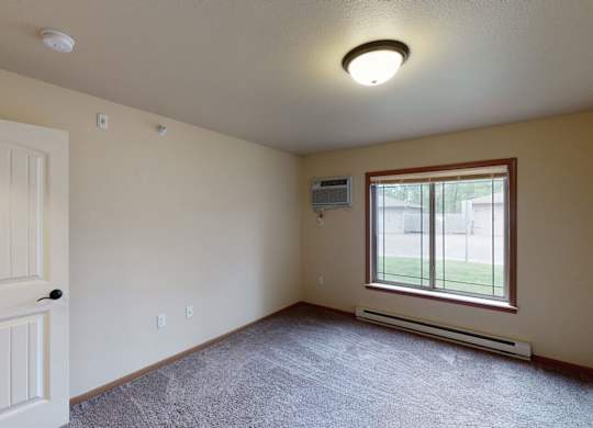A bright, empty bedroom here at Williston Garden Apartments with plush carpet, soft neutral walls, and a large window that brings in natural light and outdoor views. A wall-mounted air conditioning unit, baseboard heating, and an open layout provide comfort and flexible space for bedroom furniture.