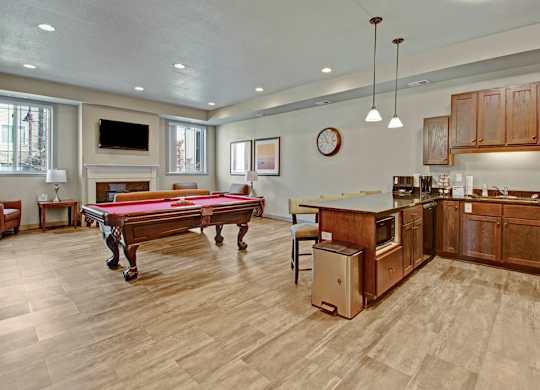 A stylish community lounge here at Williston Garden Apartments with wood-style flooring, recessed lighting, and a welcoming open layout. A pool table anchors the seating area near a wall-mounted television and fireplace, while a full kitchen with wood cabinets, an island, and pendant lighting creates a comfortable space for gatherings and social events.