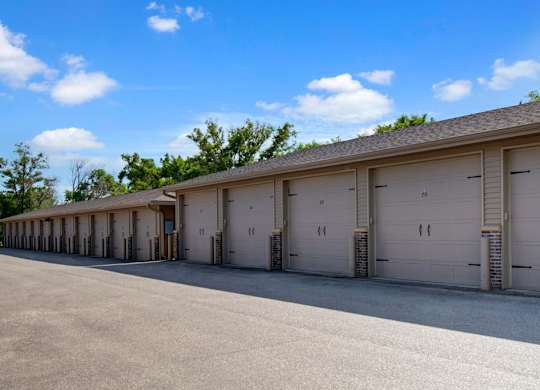 A row of private garage buildings here at Williston Garden Apartments with beige siding, white garage doors, and numbered units. The garages line a paved drive with mature trees nearby, offering convenient parking and additional storage within a clean, well-maintained community setting.