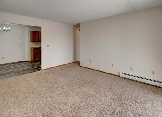 A spacious, open living area here at Windscape Apartments with soft neutral carpeting, light-colored walls, and clean baseboard trim. The room offers flexible layout options with multiple outlets along the wall and a clear connection to the adjacent dining and kitchen area, creating a bright and versatile space for everyday living.