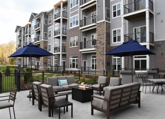 Outdoor patio of Oaks of Shorewood apartment building with lounge chairs, fire pit, and blue umbrellas. Green grass and trees in the background create a cozy vibe.