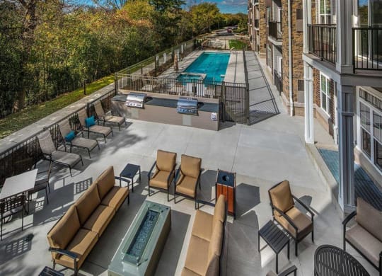 Outdoor patio at the Oaks of Shorewood Apartments with cushioned seating, a fire pit, and a barbecue area. Adjacent swimming pool with deck chairs, surrounded by trees and a sunny sky.