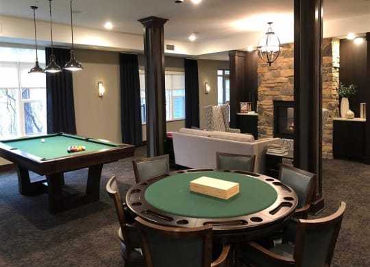 Stylish game room with a pool table and poker table at the Oaks of Shorewood Apartments. A cozy seating area by a stone fireplace exudes warmth, soft lighting, and elegance.