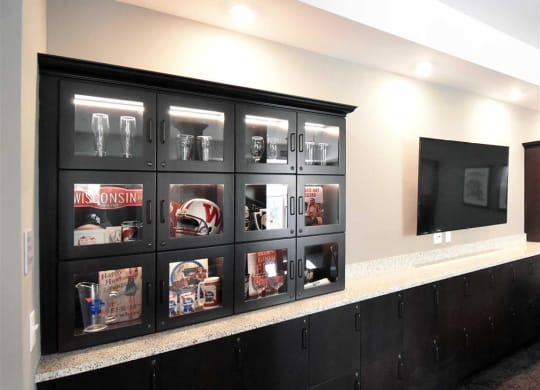 Black display cabinet with sports memorabilia and glasses at the Oaks of Shorewood Apartments, set against a light wall next to a large flat-screen TV. Modern and organized ambiance.