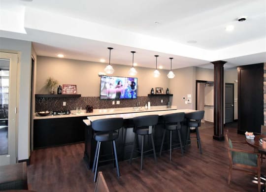 A modern lounge with a sleek bar featuring five black stools, pendant lights, and a TV displaying a sports channel, creating a relaxed, inviting atmosphere at the Oaks of Shorewood Apartments.