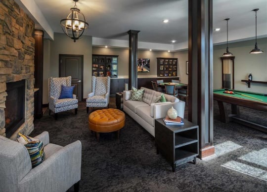 Cozy resident lounge with plush armchairs, a tan ottoman, and a beige sofa, accented with pillows. Features a stone fireplace and a pool table, creating a warm, inviting atmosphere. The Oaks of Shorewood Apartments.