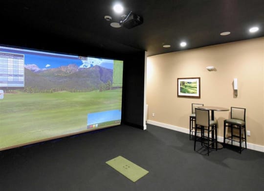 Indoor golf simulator at the Oaks of Shorewood Apartments with a large screen displaying a golf course scene. To the right, a small table with chairs and a framed picture on the wall.