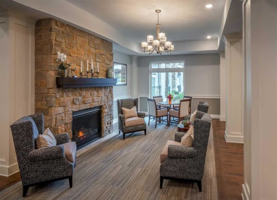 Cozy living room at the Oaks of Shorewood Apartments with a stone fireplace, flanked by cushioned armchairs. A chandelier hangs over a dining table near a large window, creating a warm ambiance.