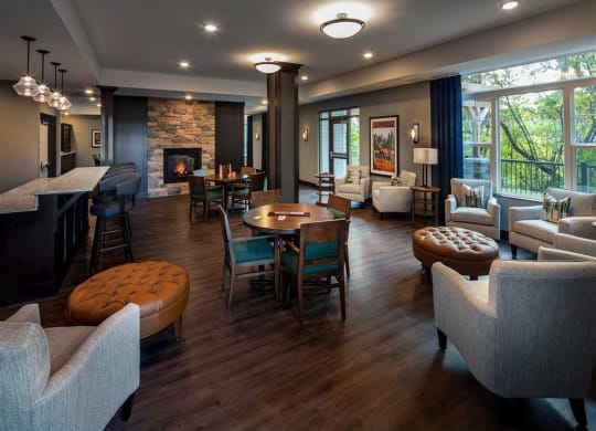 Cozy lounge at the Oaks of Shorewood Apartments with dark wood floors, plush chairs, and a stone fireplace. Large windows offer a view of greenery, creating a warm, inviting atmosphere.