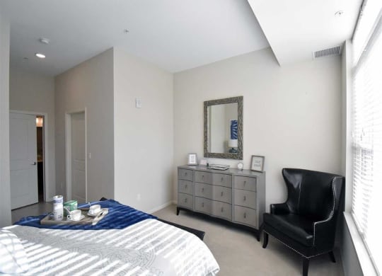 Bright bedroom at the Oaks of Shorewood Apartments with a bed, blue throw, and tray of cups. Gray dresser with mirror, black armchair, and soft lighting create a cozy, modern feel.
