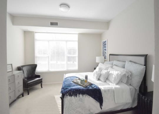 Bright bedroom at the Oaks of Shorewood Apartments with a large window, featuring a neatly made bed with a blue throw, a black armchair, and a dresser. The tone is tranquil and inviting.