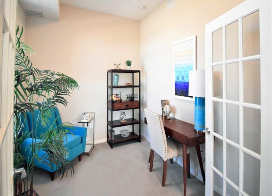 Cozy office at the Oaks of Shorewood Apartments with a blue armchair, potted plant, and shelf against cream walls. A wooden desk has a white chair, lamp, and abstract blue artwork.