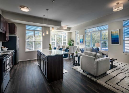 Modern open-concept kitchen and living room with dark wood floors and sleek furniture. Large windows let in natural light, creating a cozy, inviting atmosphere at the Oaks of Shorewood Apartments.