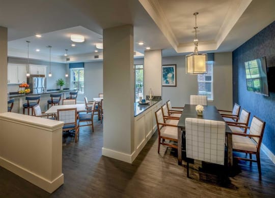 Resident lounge at the Oaks of Shorewood Apartments with modern kitchen and dining area with wood flooring, white cabinetry, and blue accents. A long dining table seats eight. Bright, welcoming atmosphere.