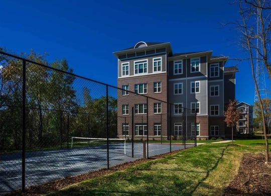 Four-story brick building with white-trimmed windows beside a fenced tennis court. Surrounded by trees under a clear blue sky, creating a peaceful atmosphere. The Oaks of Shorewood Apartments.