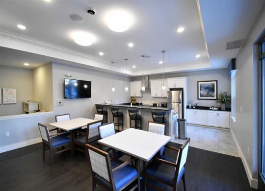 Resident lounge with modern communal kitchen with dark wood floors and gray walls at the Oaks of Shorewood Apartments. Features white cabinets, a stainless steel fridge, tables with chairs, and pendant lights.