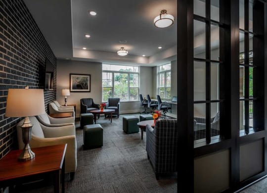 Modern lounge at the Oaks of Shorewood Apartments with soft lighting features beige armchairs, plaid chairs, and dark wood accents. Large windows provide natural light, creating a cozy atmosphere.