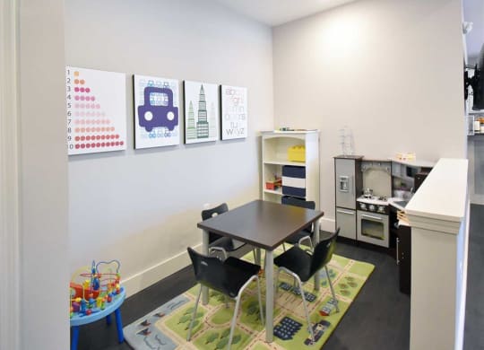 Bright playroom with a small table and four chairs on a colorful rug. Wall art shows numbers, a car, buildings, and the alphabet. Toy shelves and a pretend kitchen set give a playful vibe at the Oaks of Shorewood Apartments.