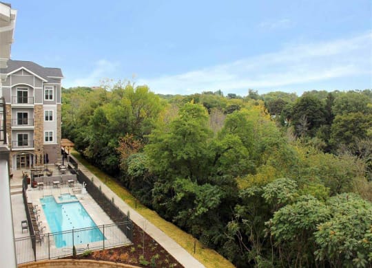 Oaks of Shorewood, a modern apartment building beside a pool with lounge chairs, bordered by lush green trees under a clear blue sky. Calm and inviting outdoor setting.