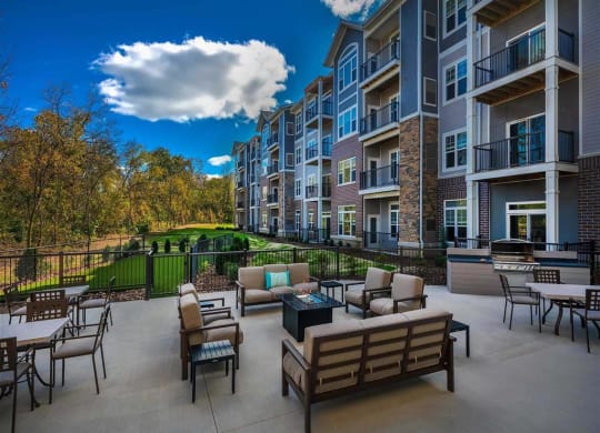 Modern, Oaks of Shorewood, apartment complex with a spacious outdoor patio featuring seating, tables, and a grill. Overlooks a grassy area with trees under a partly cloudy sky.