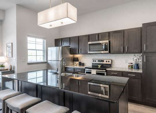 A sleek kitchen here at 98 Apartments with dark cabinetry, stainless steel appliances, glossy granite countertops, a large island with seating, and bright lighting, creating a modern and polished space for cooking and entertaining.