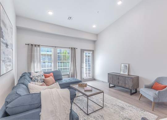 A bright living room here at 98 Apartments with wood-style flooring, a comfortable sectional sofa, soft neutral décor, and large windows with a glass door that bring in natural light, creating a calm and inviting space for everyday living.