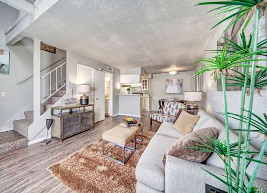 A cozy living room here at Brookwood Village with wood-style flooring, soft neutral furnishings, and warm lighting, featuring an open layout with a staircase, a small dining area, and a connected kitchen that creates an inviting and functional living space.