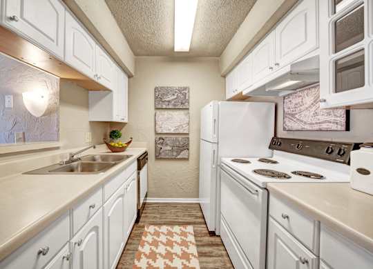 A bright kitchen here at Brookwood Village with white cabinets, light countertops, and white appliances, featuring a galley-style layout, wood-style flooring, and soft neutral accents that create a clean and welcoming cooking space.