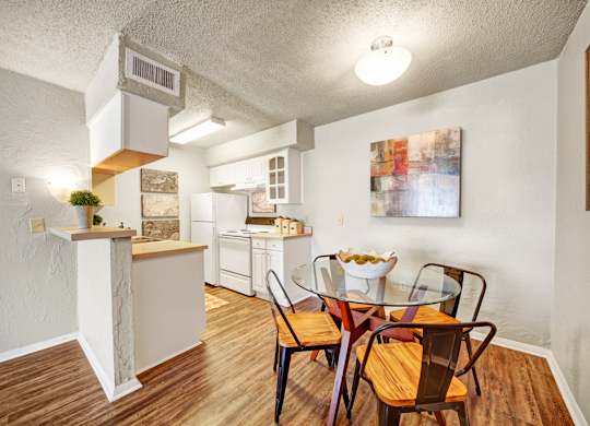 A charming dining area here at Brookwood Village with wood-style flooring, a round glass-top table, and warm accent lighting, featuring an open view into the kitchen that creates a comfortable and connected dining space.