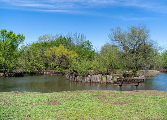 A bench sits on a grassy area next to a body of water with trees in the background.