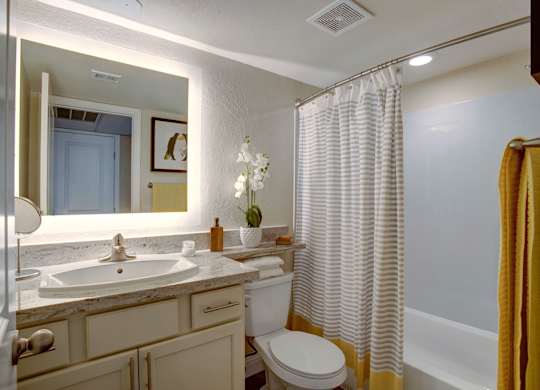 A clean bathroom here at Indigo with a bathtub and shower combo, light countertops, and white cabinetry. A large illuminated mirror and neutral finishes create a bright, fresh, and comfortable space.